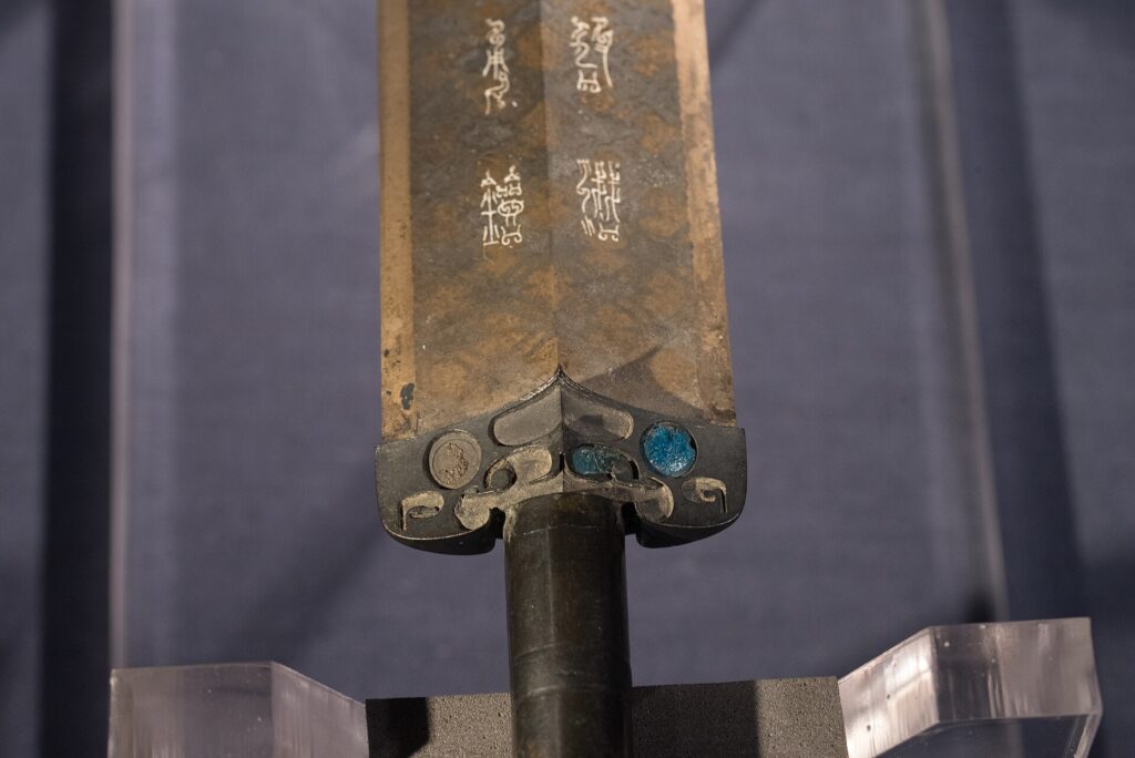 越王勾践剣の鍔の近く。鳥の形を含む8文字の銘と、鍔にはめ込まれた青い玉が見える