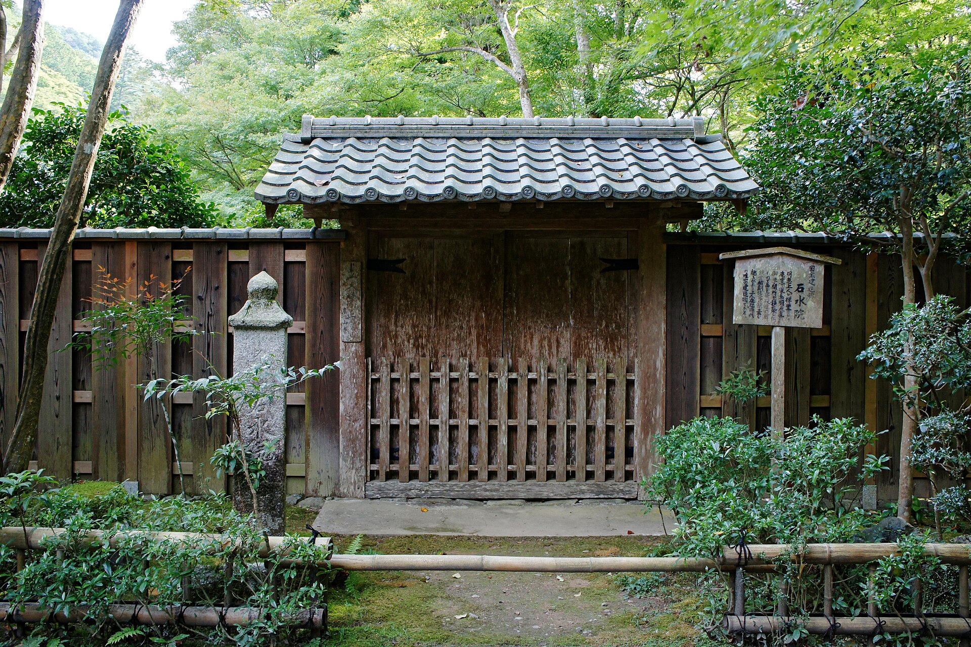 石水院の門。板塀と瓦屋根の小さな門の脇に石灯籠と説明の札が立つ